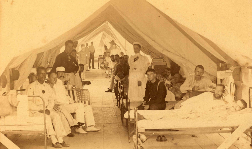Sepia photo of a field hospital tent with patients lying on beds and nurses and staff standing and sitting throughout the tent.
