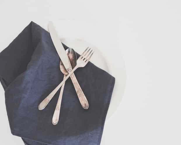 A white plate with a dark napkin placed on it, topped with a knife, spoon, and fork arranged in a triangular pattern.