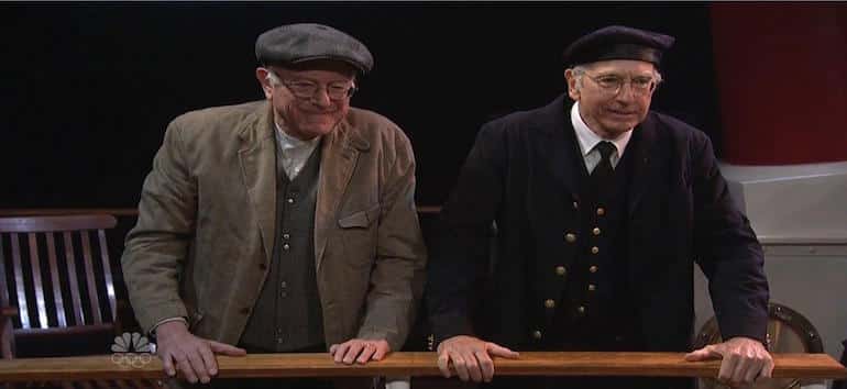 Two older men in vintage-style clothing stand on a ship deck, each wearing a flat cap and a coat.