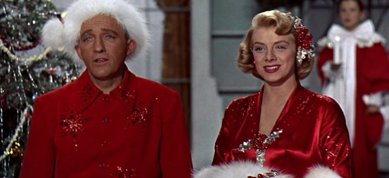 Two people dressed in festive red outfits and white fur-trimmed hats stand in front of a decorated Christmas tree and another person dressed in a red robe stands in the background.