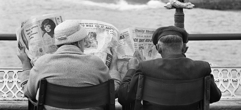 elderly coupe sitting outdoors reading the newspaper