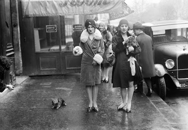 Two women in vintage fashion walk with a small dog on a wet street, surrounded by other pedestrians and parked cars, in front of a shop with signs in the windows.