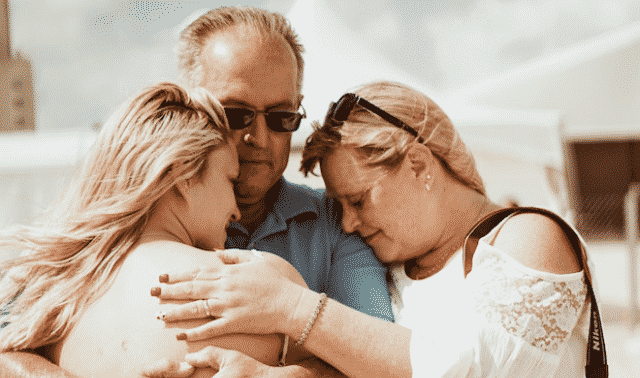 Three people are hugging. The person on the left has blond hair and is facing the person in the center, who is wearing glasses. The person on the right also has blond hair and is leaning in for the hug.