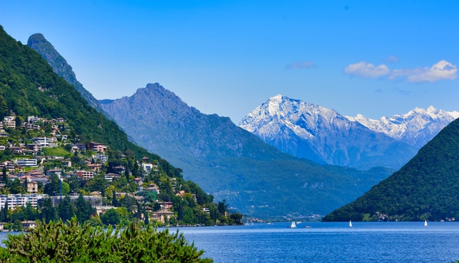 A lake surrounded by green hills and snow-capped mountains, with a few boats on the water and houses on the hillside.