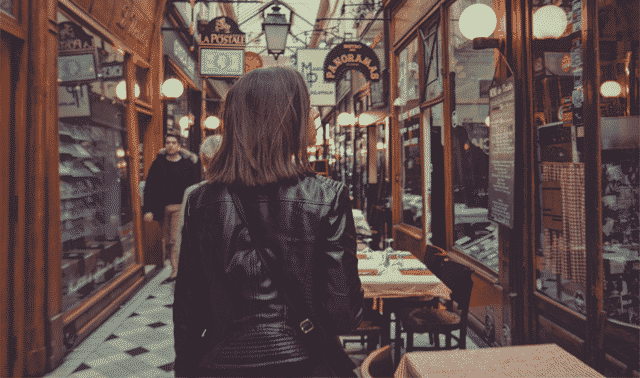 A person in a black leather jacket walking through a narrow, dimly lit market or arcade with shops and cafes on both sides. Several illuminated signs and lanterns are visible.