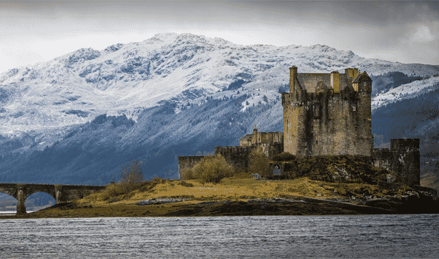 A medieval castle sits on a small island with a stone bridge connecting to the mainland, surrounded by a lake and snow-capped mountains in the background.