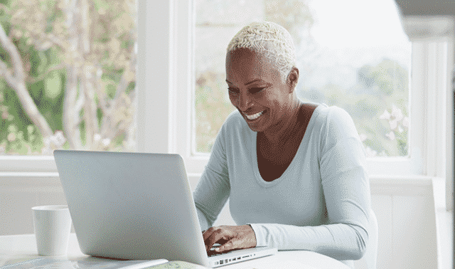 A person with short gray hair smiles while using a laptop at a bright, airy table with a white mug nearby.