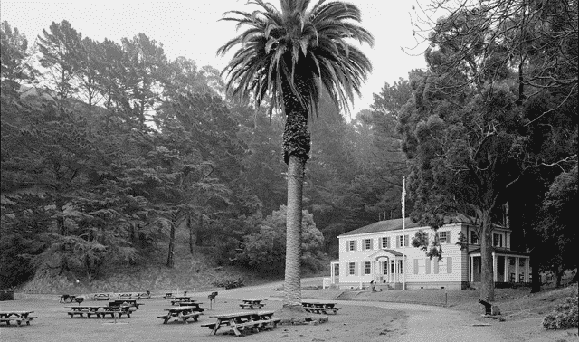 A two-story building stands near a tall palm tree, surrounded by picnic tables and trees, with a forested hillside in the background.