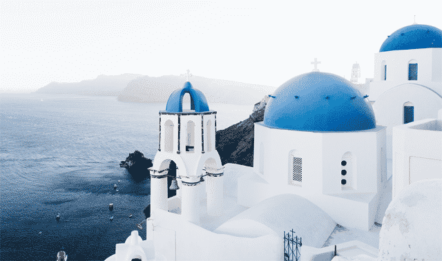 White buildings with blue domes overlook the sea in Santorini, Greece, with mountains visible in the background.