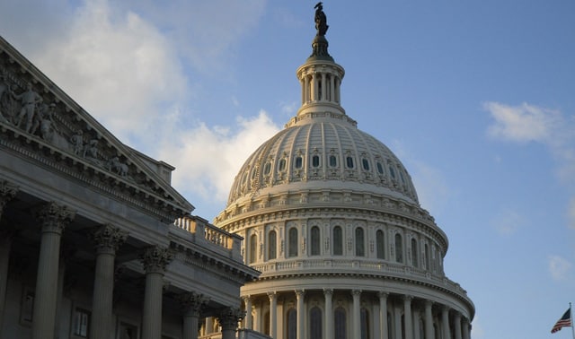 The dome of the United States Capitol Building against a blue sky. The building features neoclassical architecture with decorative sculptures visible on the exterior.