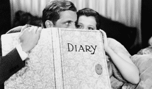 A man and woman peek over a large book labeled "DIARY" in a black-and-white image.