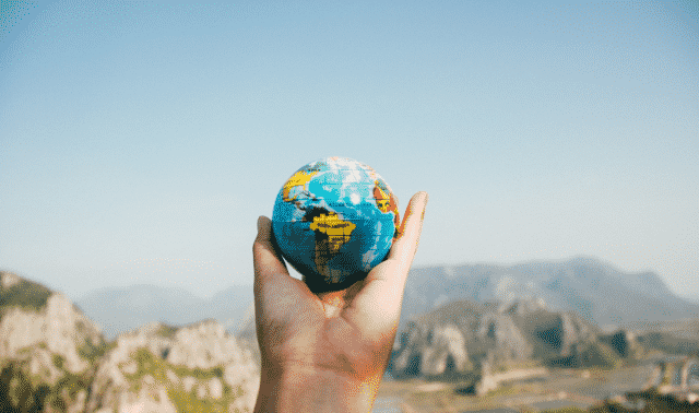 A hand holding a small globe with a mountainous landscape in the background under a clear blue sky.