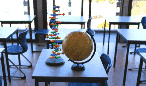 A classroom with desks and chairs, featuring a model of a DNA double helix and a globe placed on a table near the windows.