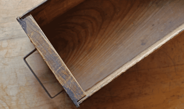 Photo of an empty wooden drawer with a metal handle, shown from an angle that highlights the wood grain and worn edges.