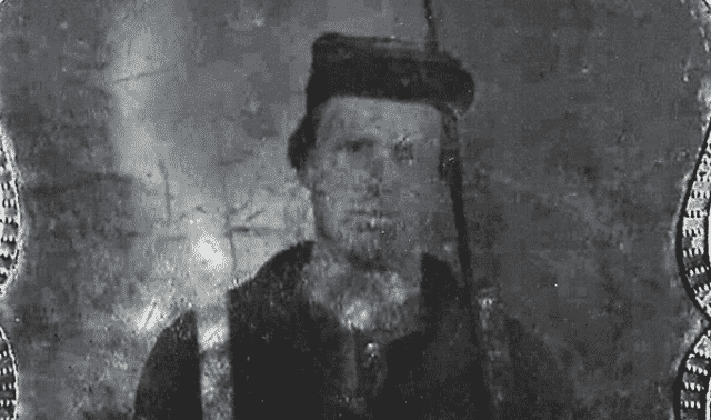 A black-and-white photograph of a man wearing a dark coat and a cap. The image appears aged and grainy, indicating it is an old photo.