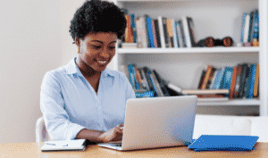 A person sits at a desk using a laptop, with a notebook and blue folder nearby. Bookshelves filled with books are in the background.