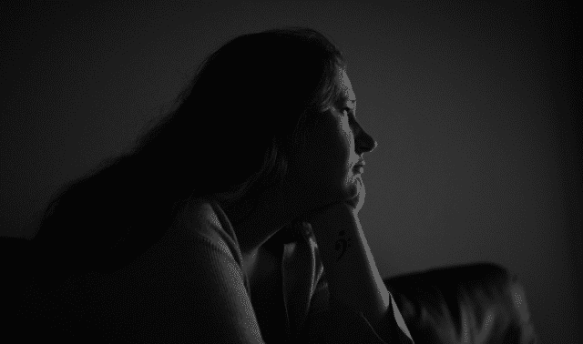 A person in a dimly lit room rests their chin on their hand, gazing thoughtfully into the distance.