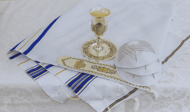 A gold goblet with a saucer, a white fringed prayer shawl with blue and gold stripes, an embroidered belt, and three white kippahs are displayed on a white cloth-covered surface.