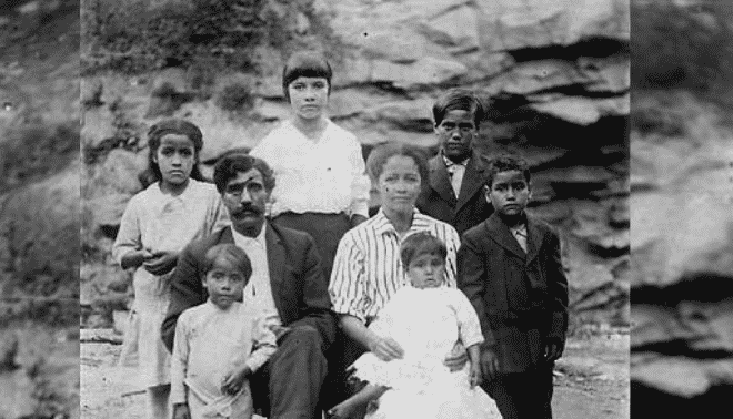 A black and white photograph of a family with eight members. Four children stand behind their seated parents, while a young child sits on the mother's lap and another stands beside the father.