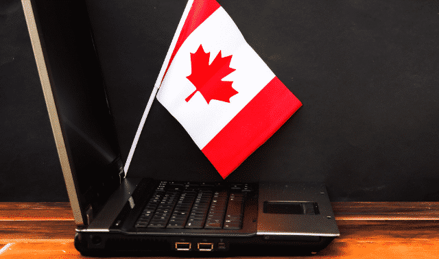 A small Canadian flag is placed inside the keyboard of an open laptop on a wooden surface against a dark background.