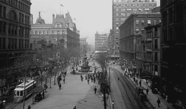 Black and white image of a busy city street from the early 20th century with a fountain, buildings, people walking, and a streetcar on the left.