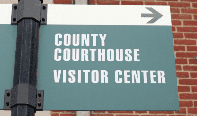 A sign reading "County Courthouse Visitor Center" with an arrow pointing to the right is mounted on a black post against a brick wall backdrop.