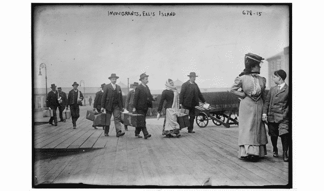 Black and white photo of immigrants arriving at Ellis Island, carrying suitcases and walking on a wooden dock.