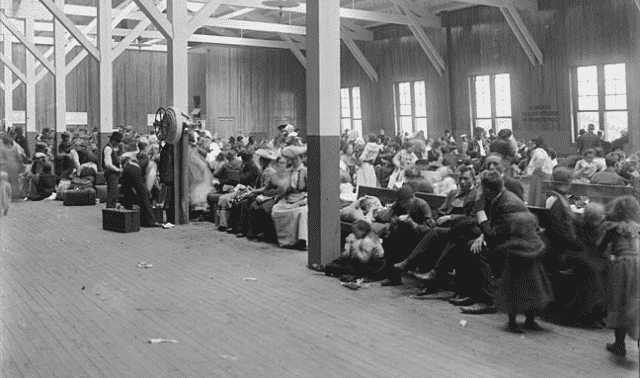 A crowded room of people, many sitting and standing, inside a large wooden building with columns and high ceilings.