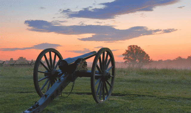 A historic cannon sits on a grassy field at sunrise, with a misty atmosphere and trees in the background.