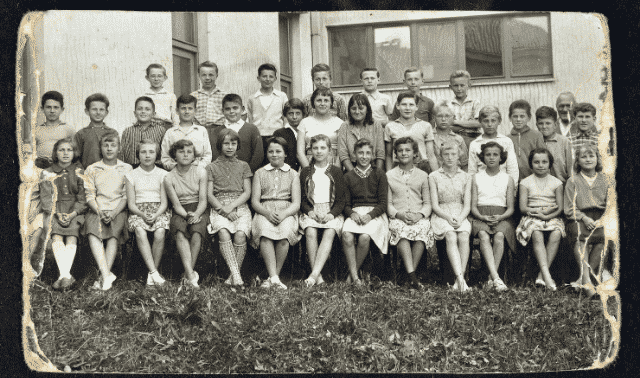 A black-and-white aged photo shows a group of 36 children in four rows posing for a school class picture in front of a building.