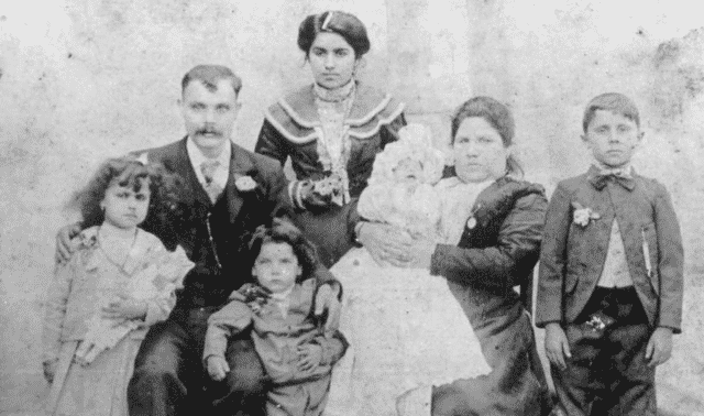 A vintage black and white photograph featuring a family of seven posing for a portrait. Two women, two men, and three children, dressed in early 20th-century attire, stand and sit together.