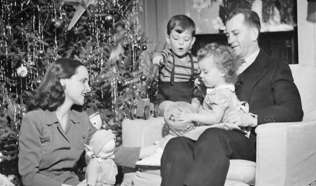 Family opening presents near Christmas tree, featuring a father, mother, young boy, and young girl