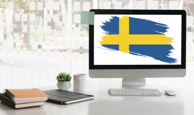 Computer monitor displaying the Swedish flag on a desk with a keyboard, notebook, and plant in an office setting.