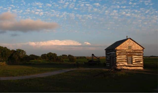 A small wooden log cabin sits on a grassy field under a sky with scattered clouds, with trees and farmland in the background.