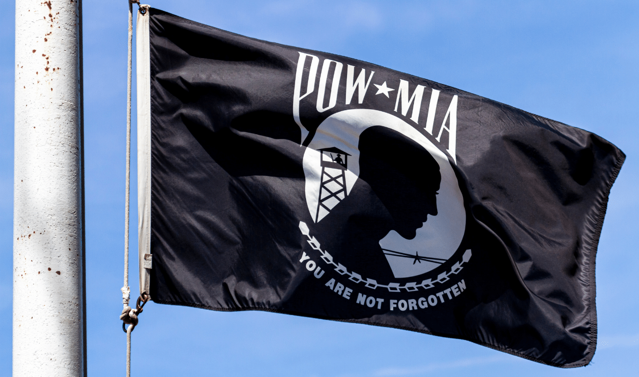 A black POW*MIA flag waves on a flagpole against a blue sky, displaying the silhouette of a man, guard tower, barbed wire, and the words "You Are Not Forgotten.