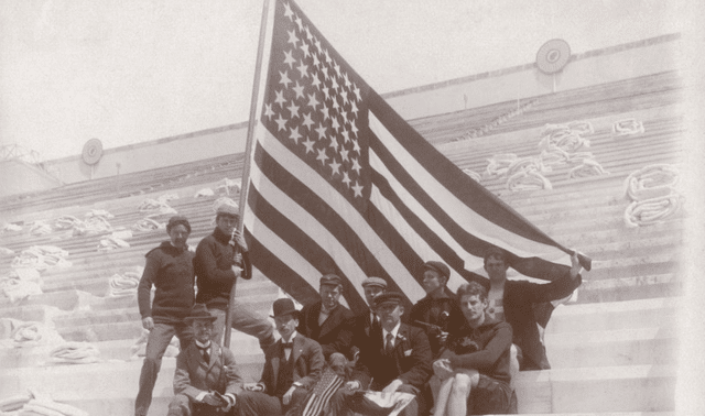 A group of men sit and stand on stadium steps holding a large American flag; empty seats with coiled ropes are in the background.