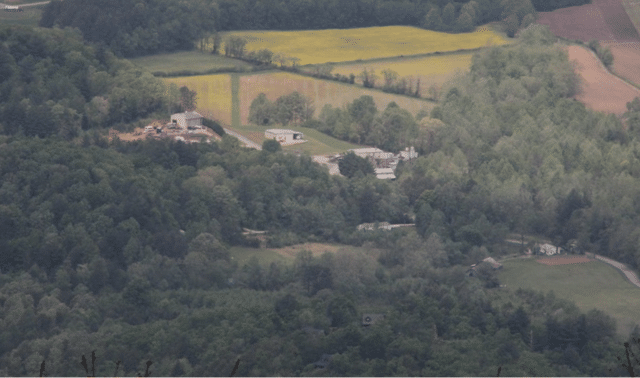Aerial view of a rural area with scattered buildings surrounded by dense green trees and fields, with a winding road visible in the lower right corner.