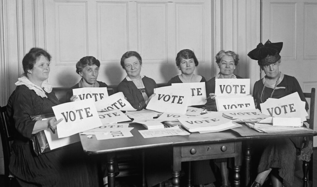 Six women sitting at a table hold and display "VOTE" signs, surrounded by more signs and papers, in a formal indoor setting.