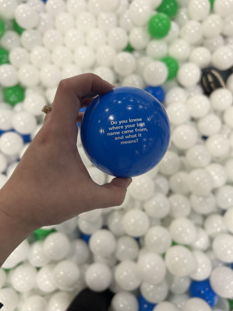 A hand holds a blue plastic ball with the words, "Do you know where your last name came from, and what it means?" above a pit filled with white, green, and blue balls.