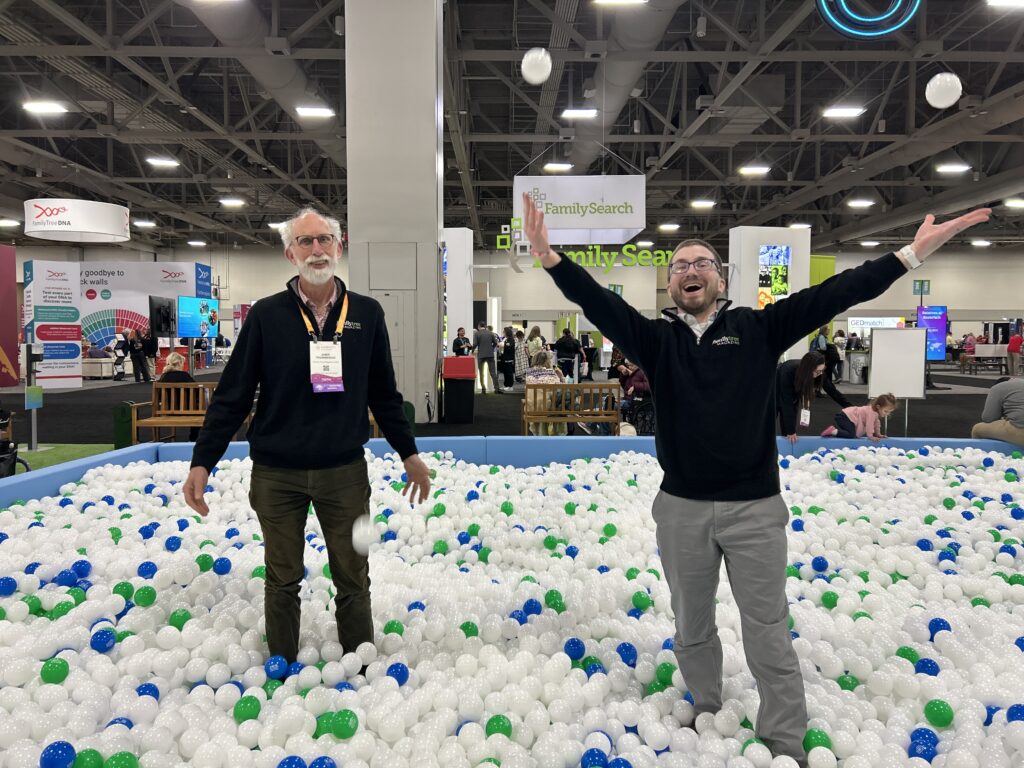 Two men stand in a large ball pit at an indoor event; one man is tossing balls in the air while the other stands still. Booths and banners are visible in the background.