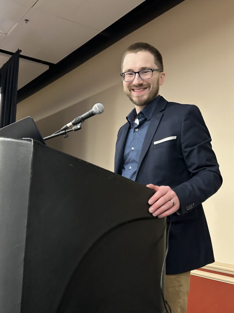 A man wearing glasses and a navy blazer stands at a podium with a microphone, smiling at the camera in an indoor setting.