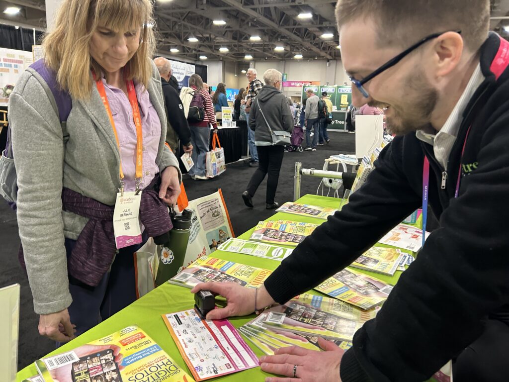 A man stamps a brochure while a woman watches at a booth covered with magazines during a busy convention or trade show.