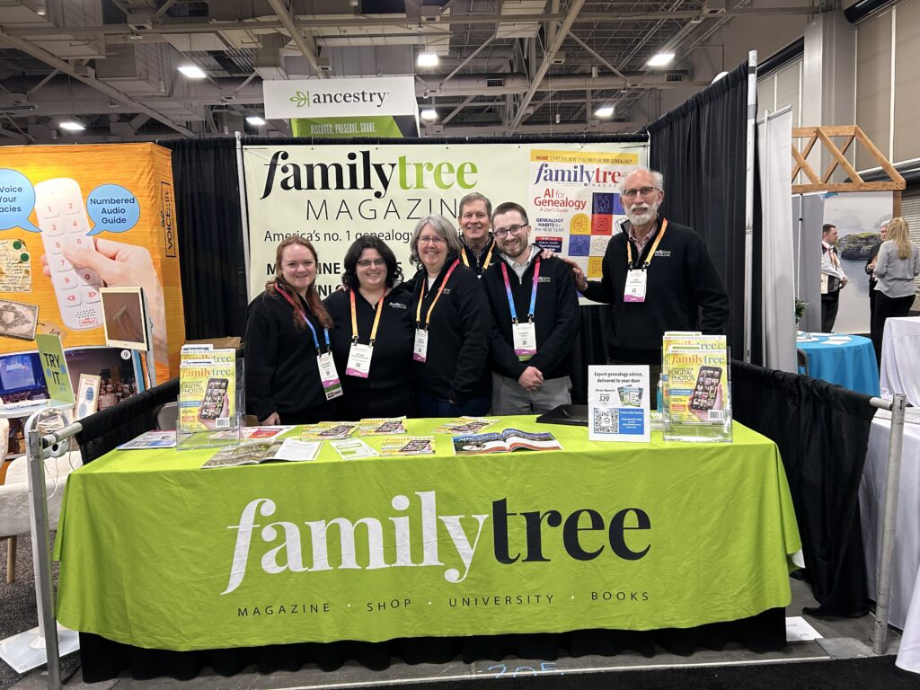 Six people stand behind a table covered with Family Tree Magazine materials at a genealogy conference booth with an Ancestry banner overhead.