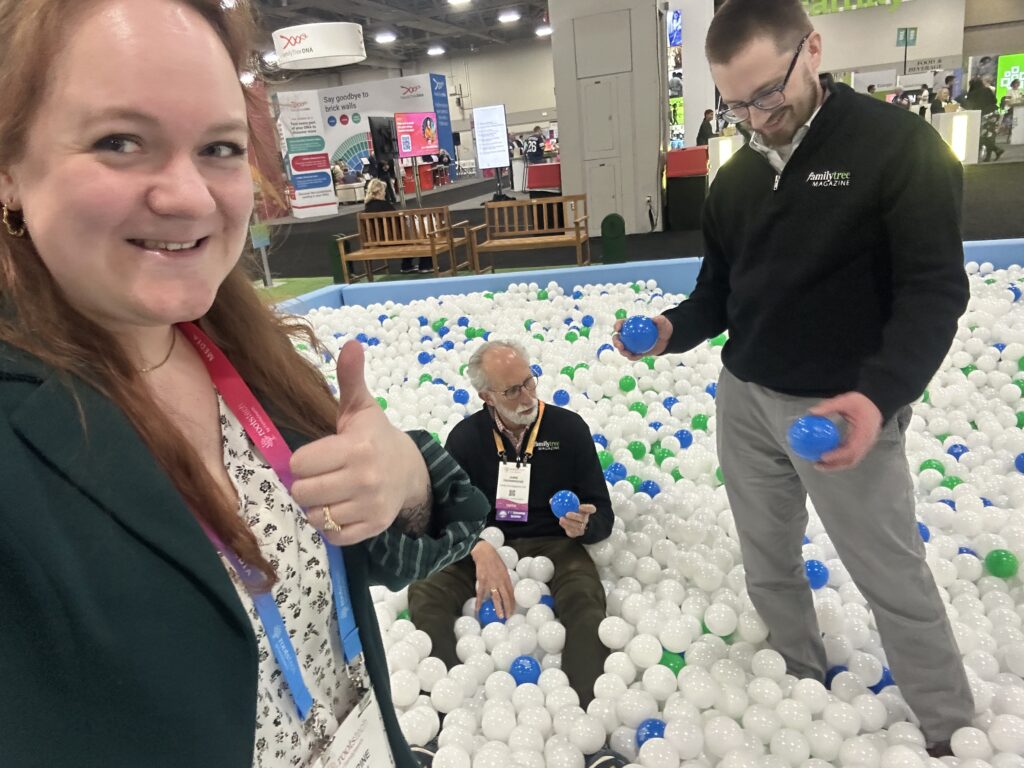 Three adults in business attire are smiling and interacting inside a ball pit at a convention or trade show.