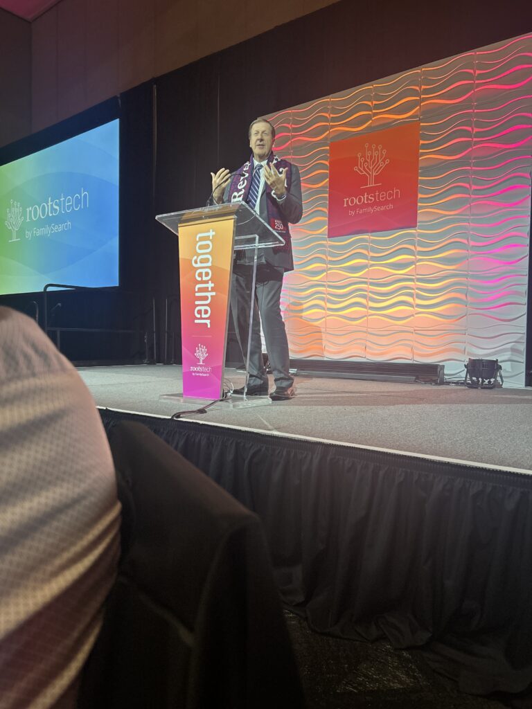 A speaker stands at a clear podium on stage at RootsTech, with colorful event signage and a wavy backdrop behind him.