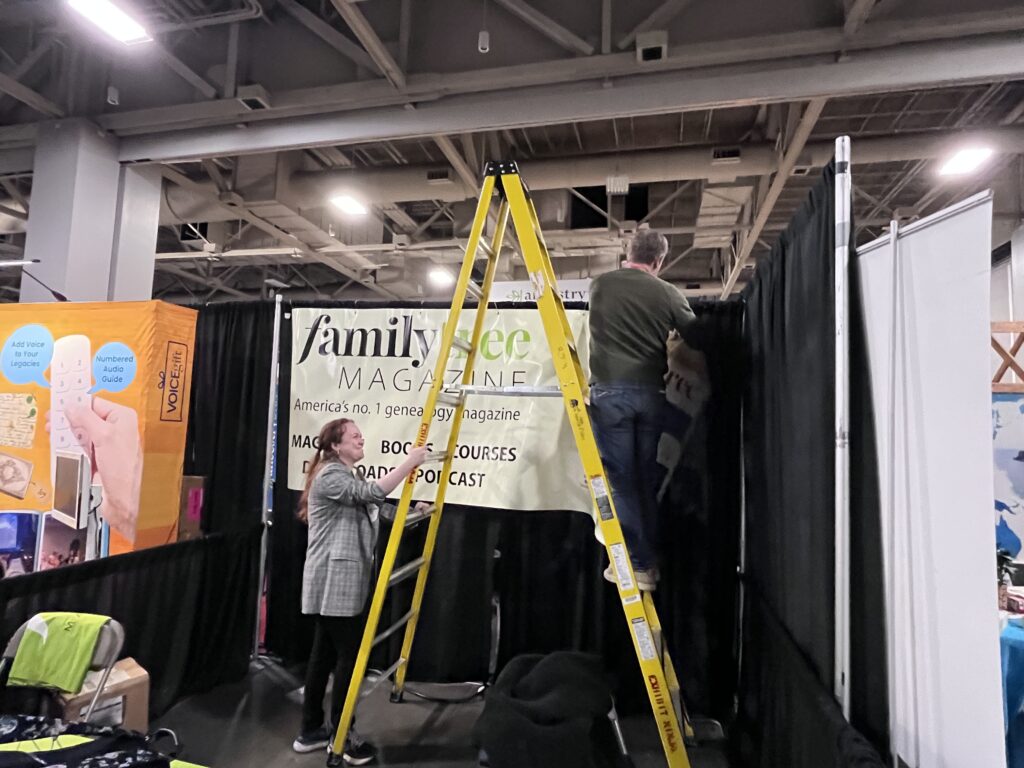 Two people set up a booth for Family Tree Magazine at an indoor event, with one person on a ladder hanging a banner and another person assisting from the ground.
