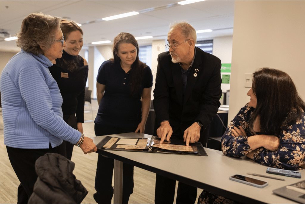 Five people stand or sit around a table, closely examining documents together in a brightly lit office setting.