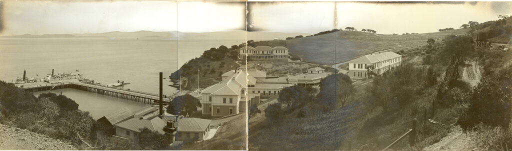 Historic photograph of a coastal facility with multiple buildings on a hillside, a dock extending into the water, and distant mountains visible across the bay.