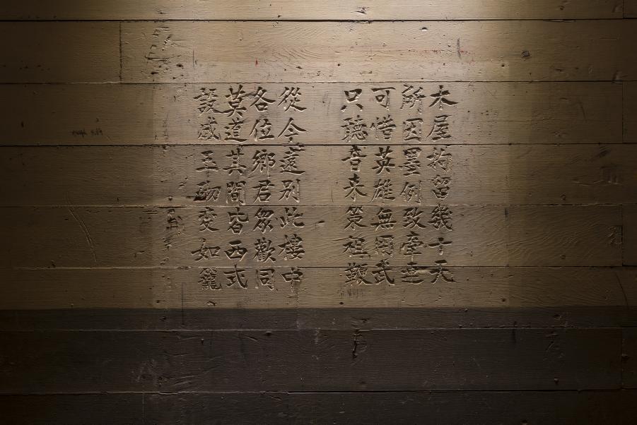 Chinese characters are handwritten in vertical columns on a wooden wall with a beam of light illuminating the center.