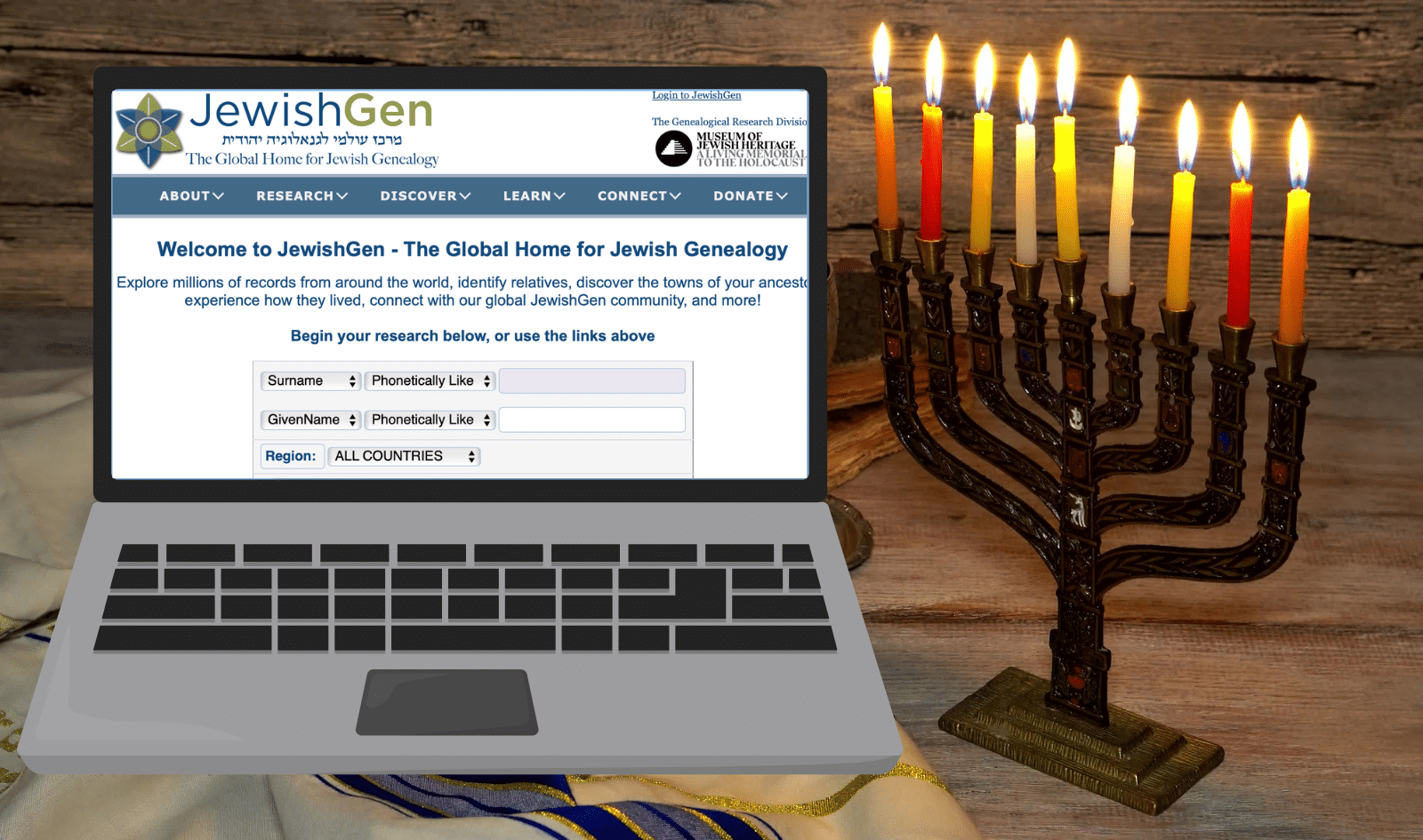 A laptop displaying the JewishGen genealogy website sits next to a lit menorah with eight candles on a wooden surface.
